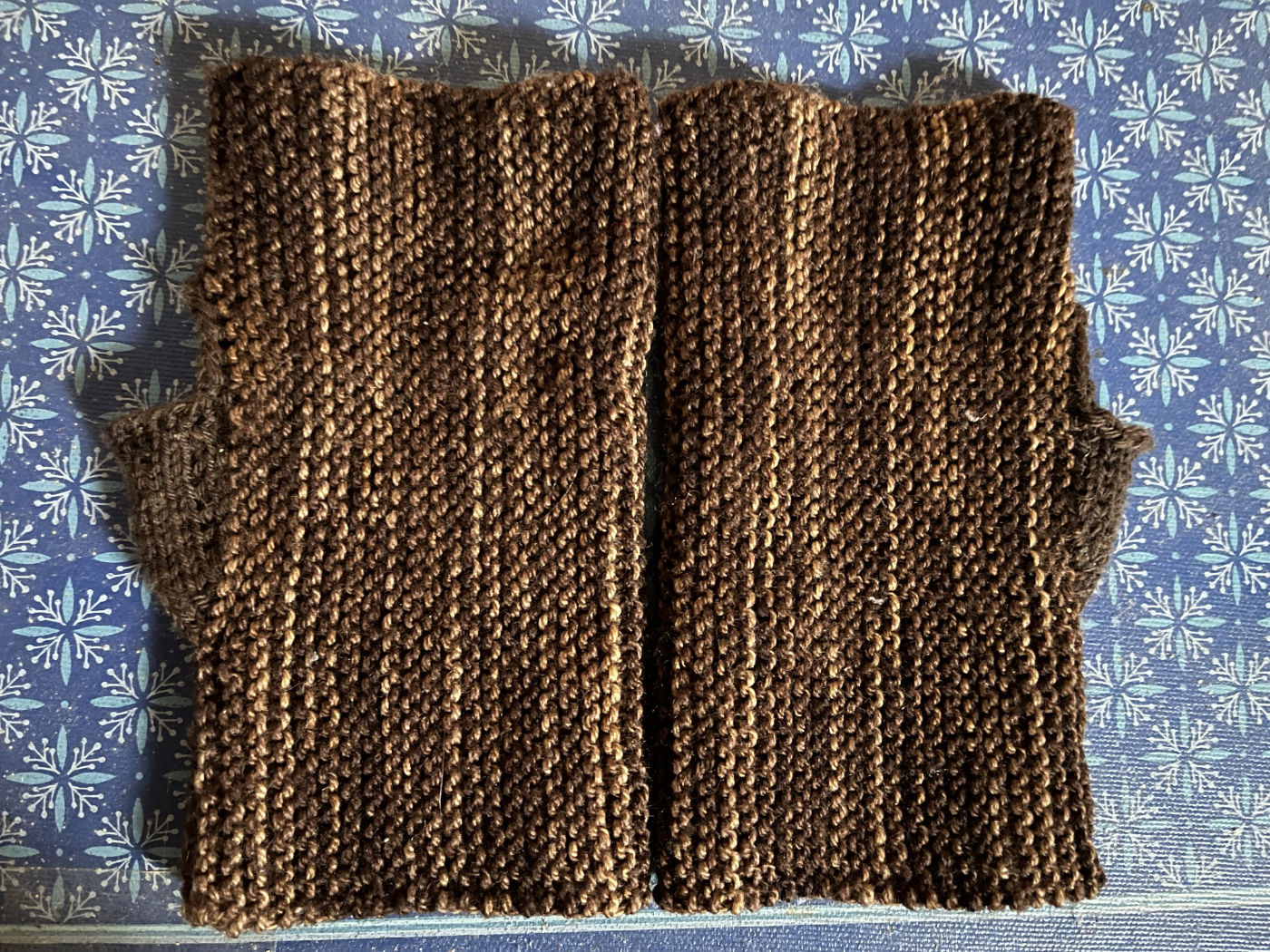 The pair of brown mitts sitting flat on a blue book. They're two flat tubes, each with one shallow brown triangle sticking out to one side. This covers the base of the thumb when worn.